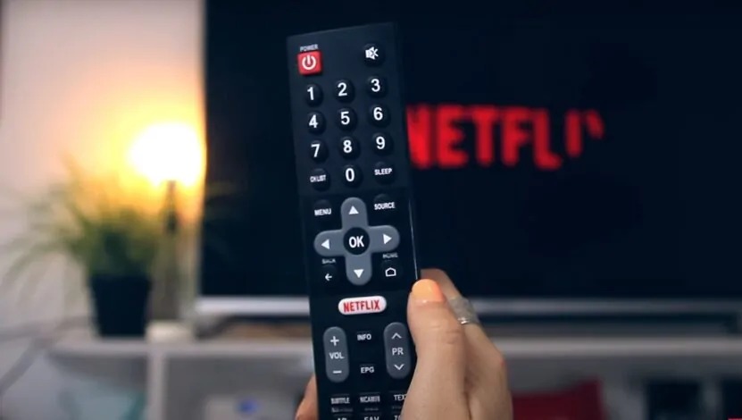 Hand holding a TV remote pointed at a screen displaying the Netflix logo