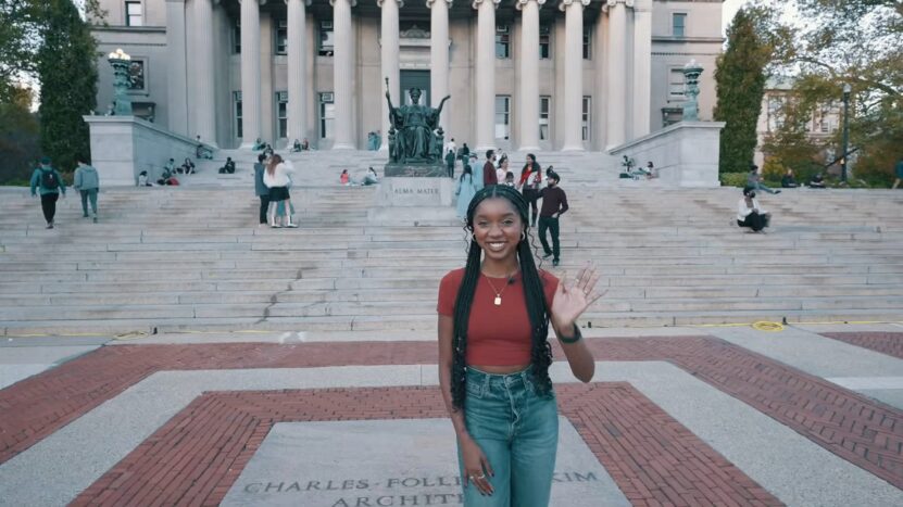 A young woman with braided hair, wearing a red crop top and jeans, waves in front of a grand university building with tall columns and the Alma Mater statue, while students walk and sit on the steps behind her