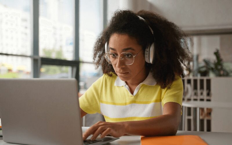 Young woman wearing headphones and glasses focused on researching school information on a laptop.