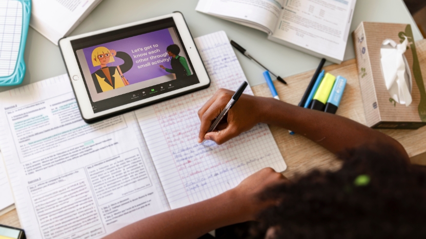 A student attending a hybrid learning session on a tablet while taking notes in a notebook
