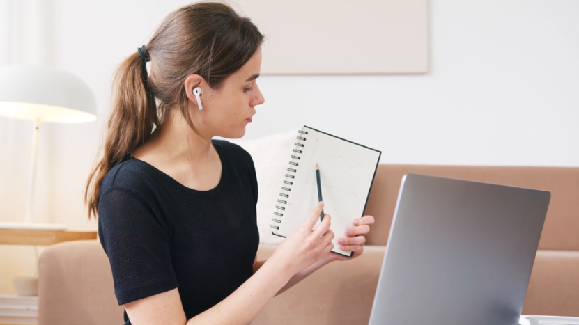 A student participating in hybrid learning, using a laptop and taking notes while wearing wireless earbuds