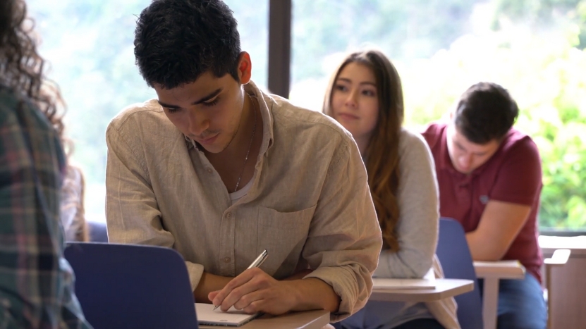 A Student Focuses on Taking Notes in Class While Others Are Seated in The Background