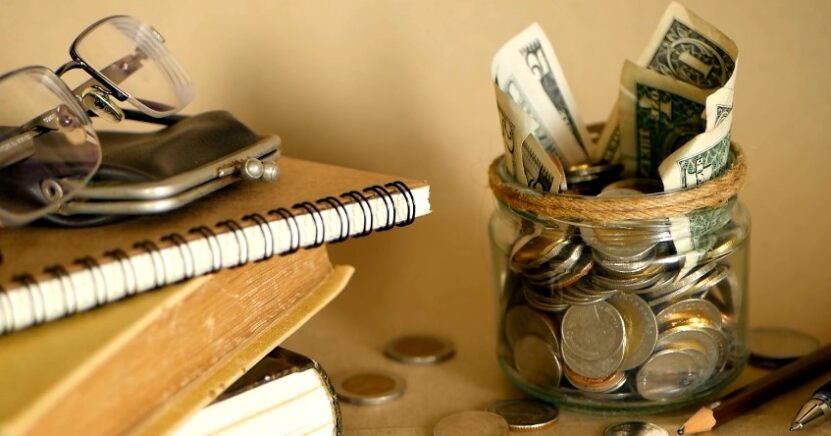A glass jar filled with coins and bills, nestled amongst books and eyeglasses