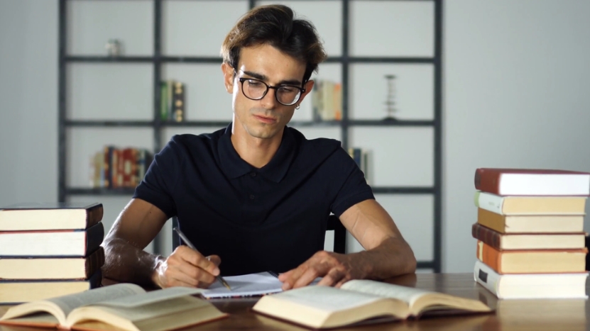 A student focuses on his studies, writing notes while surrounded by books