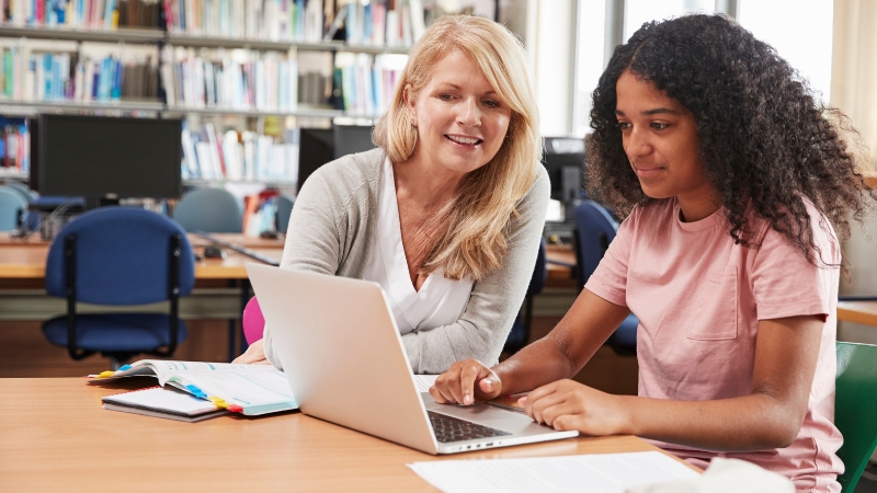 A teacher and student work together on a laptop in a library, using Google Workspace for classwork and support