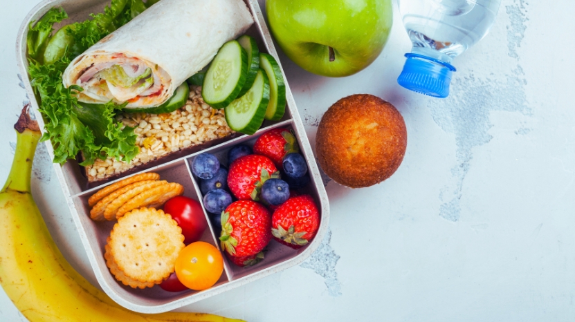 A balanced emergency school meal with fresh fruit, crackers, a wrap, bottled water, and a muffin arranged in a lunch container