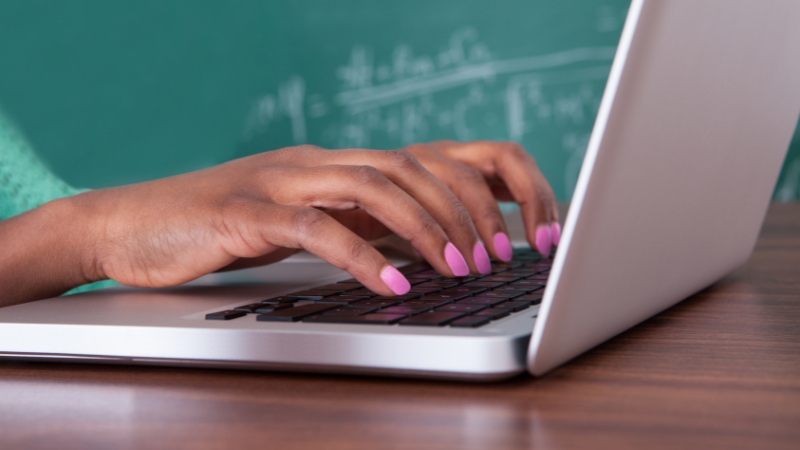 Close-up of a student typing on a laptop in front of a chalkboard, using digital tools for learning