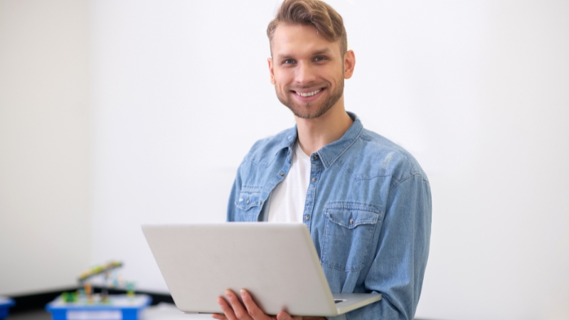 Smiling teacher holds a laptop while standing in a classroom, ready to use Google Workspace tools