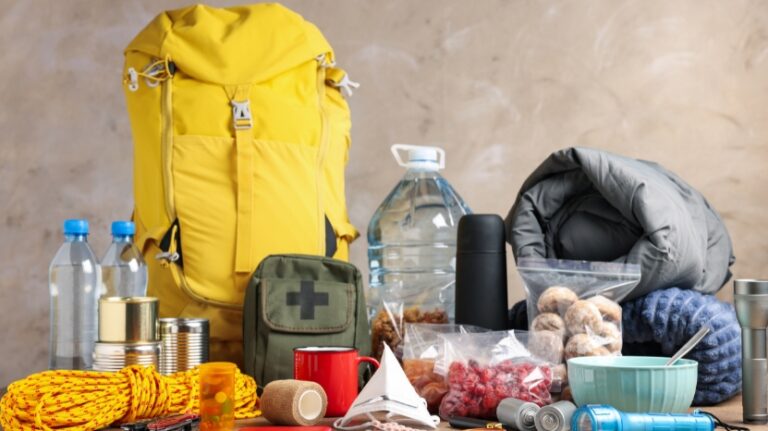 Emergency supplies including water, food, first aid kit, flashlight, rope, and sleeping bag arranged with a yellow backpack
