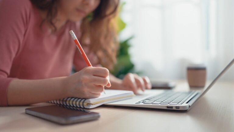 A young woman is studying alone at a desk, taking notes in a notebook while using a laptop