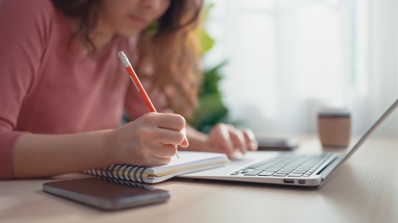 A young woman is studying alone at a desk, taking notes in a notebook while using a laptop