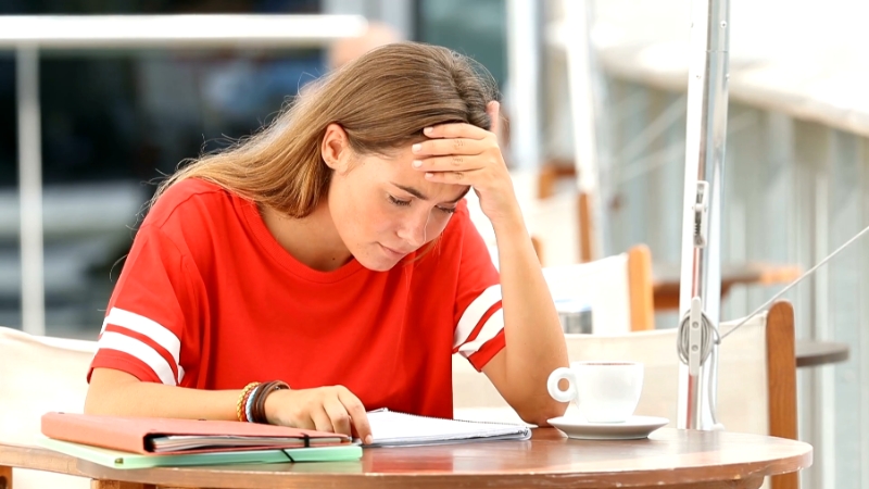 A student sits alone at a table, looking frustrated and holding her head while studying without asking for help
