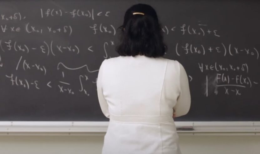 A woman in a white coat stands before a blackboard filled with mathematical formulas