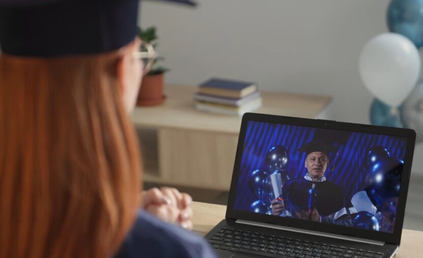 A woman in a graduation gown watches a video on her laptop, celebrating her academic achievement