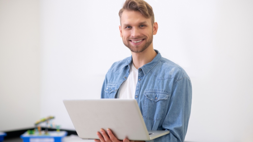A smiling man holds a laptop, ready to plan a quiz before creation in a bright classroom