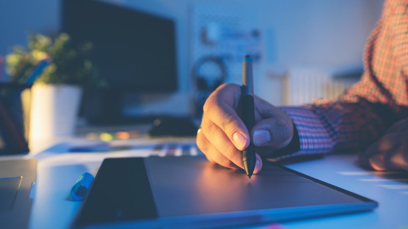 A person uses a digital drawing tablet and pen to create a design at a desk with a computer in the background