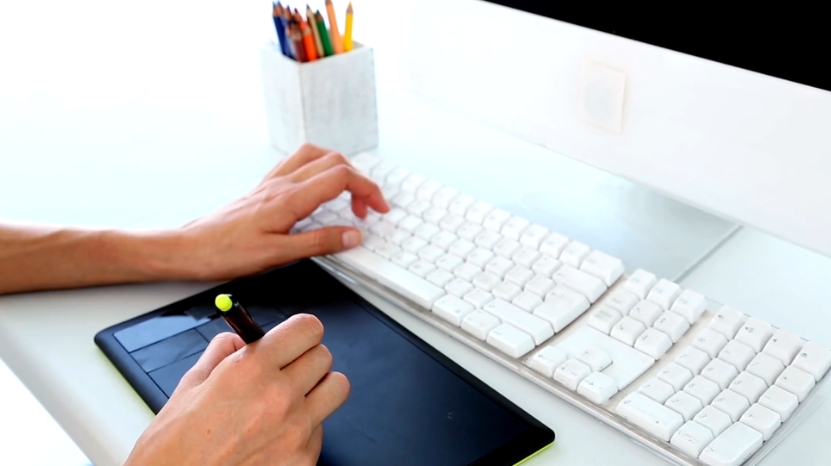 A person uses a drawing tablet and keyboard at a clean desk to work on graphic design projects