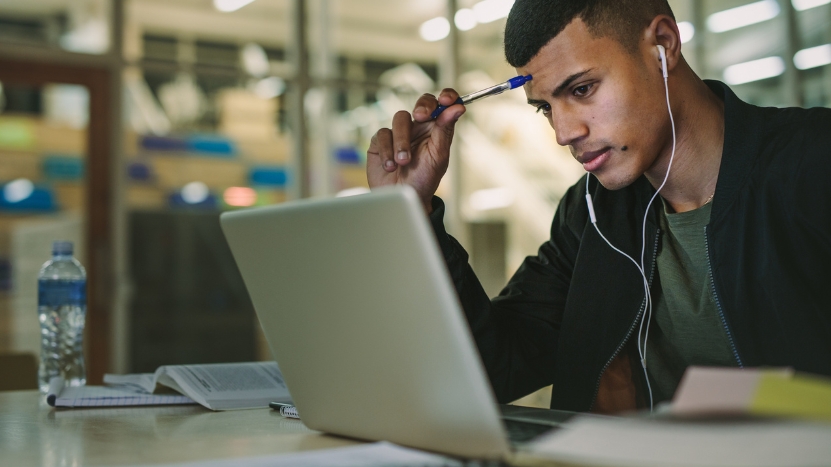 A focused student wears earphones and studies on a laptop at a campus library table with notes and a water bottle nearby