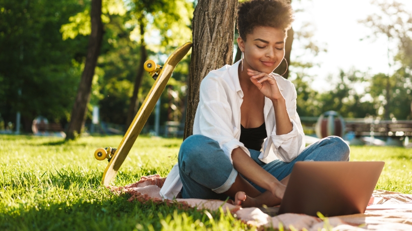 A student sits barefoot on a blanket in the grass, using a laptop under a tree with a skateboard resting nearby