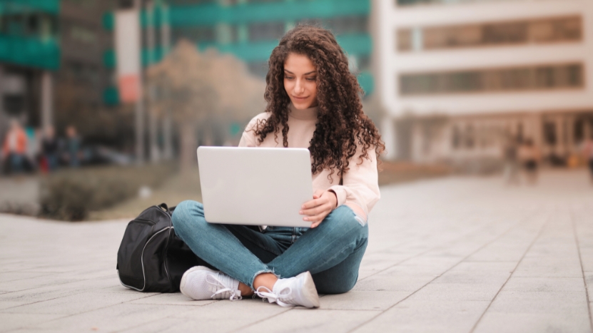 A college student sits cross-legged on campus using a laptop, with a backpack beside her