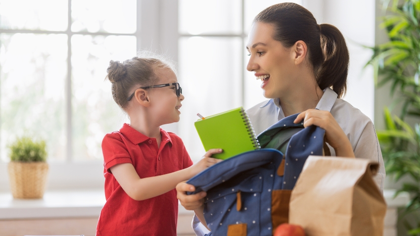 A mother helps her daughter pack a backpack with school supplies while smiling together in the morning
