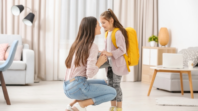 A mother kneels to check on her daughter with a yellow backpack before school in the morning