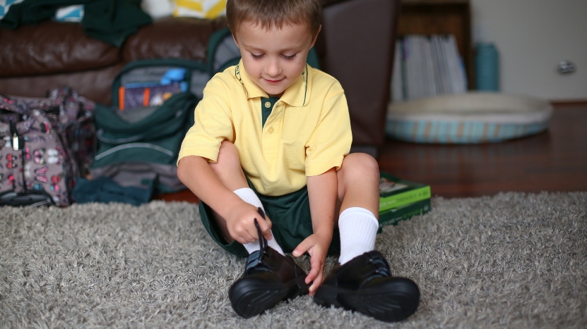 A young boy in a school uniform puts on his black shoes while sitting on the carpet