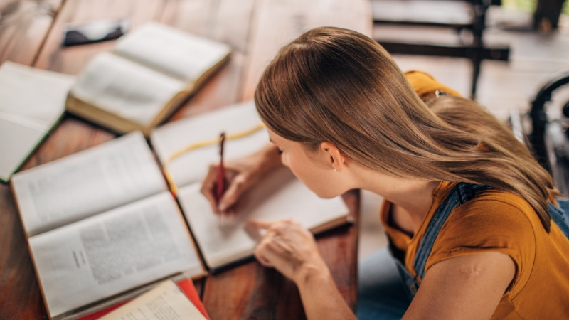 A student writes notes while studying with open textbooks spread out on the table