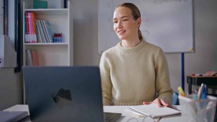 A teacher sits at a desk with a laptop and notebook