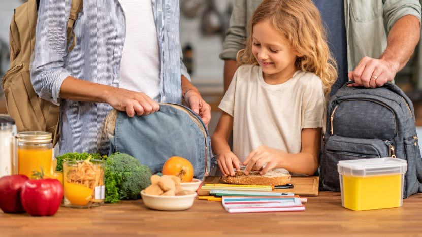 A young girl prepares a sandwich with her parents while packing school bags and snacks the night before