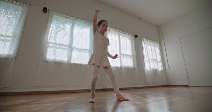 A young girl practicing ballet moves in a dance studio with large windows