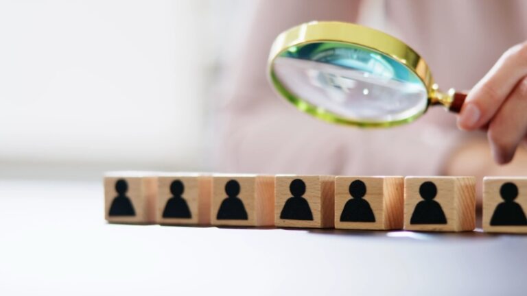 A magnifying glass inspects wooden blocks with people icons for fractional executive hiring